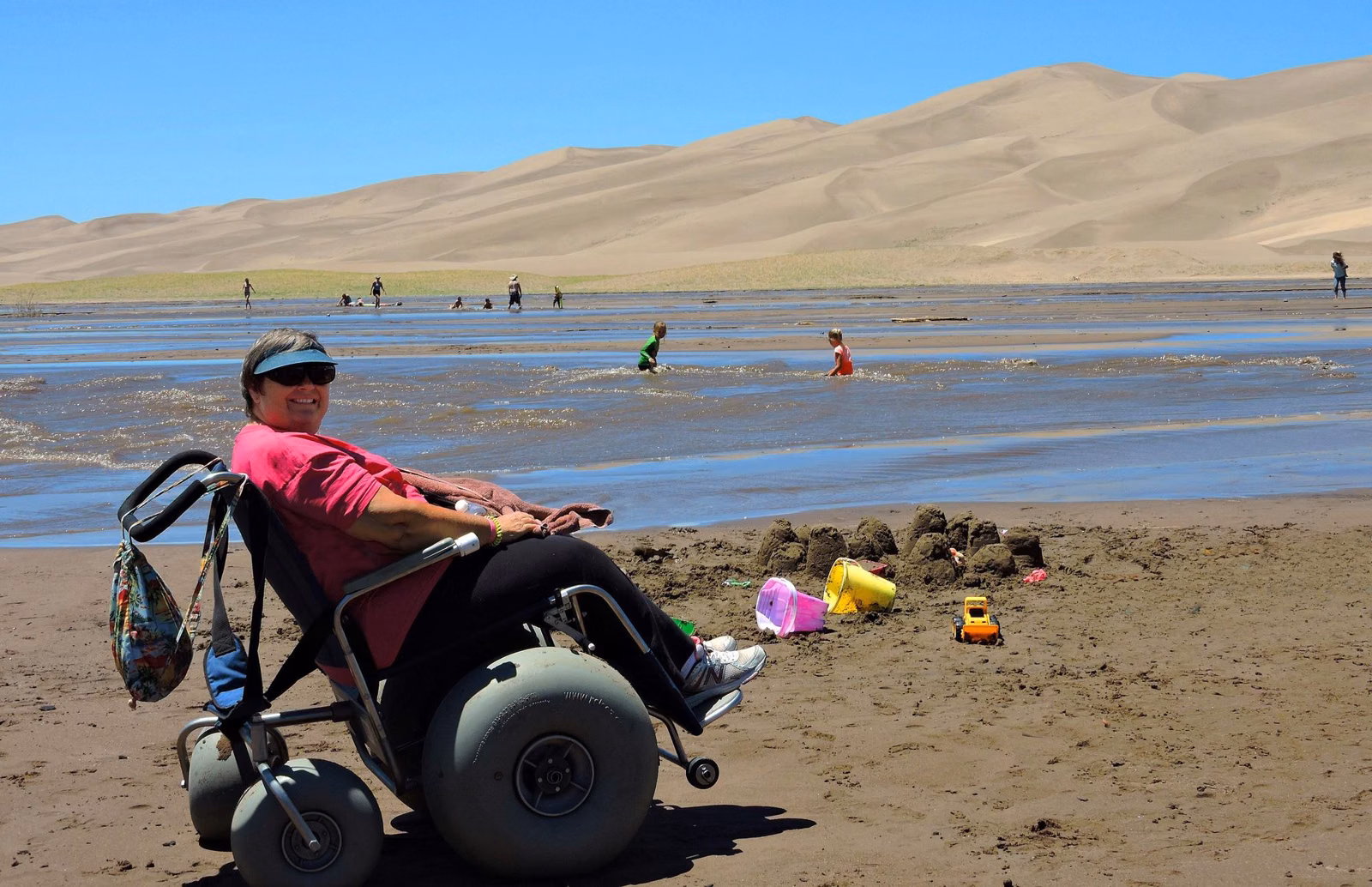 A visitor in a beach wheelchair with balloon tires on wet sand by shallow water