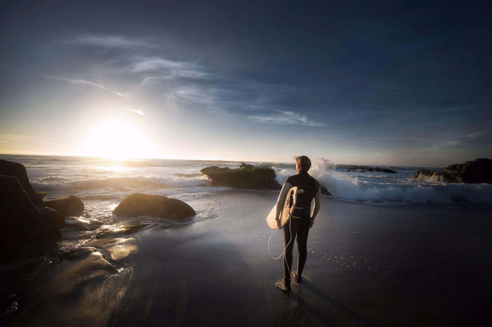 A surfer with a board walking along a rocky shore at sunrise