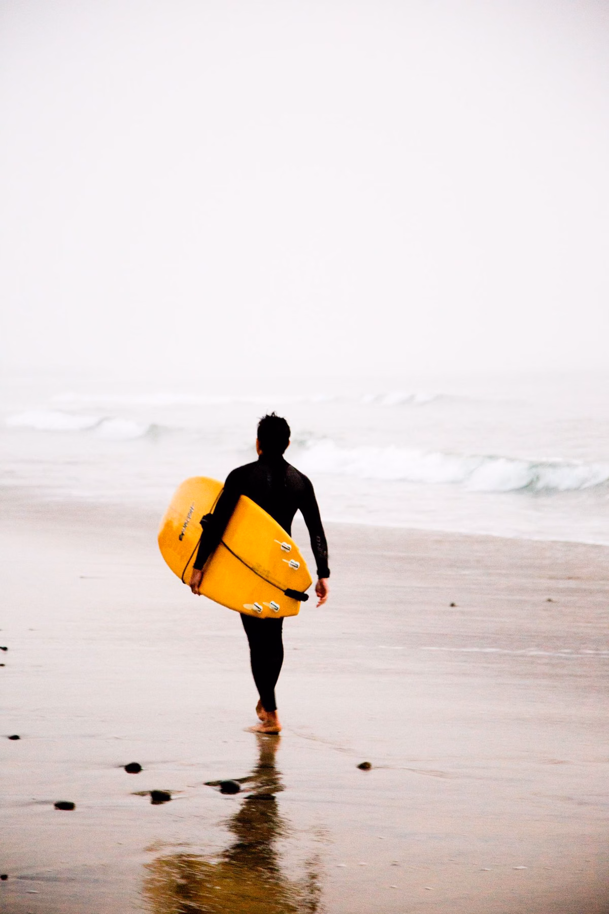 A surfer in a wetsuit carrying a yellow board toward the water in Carlsbad